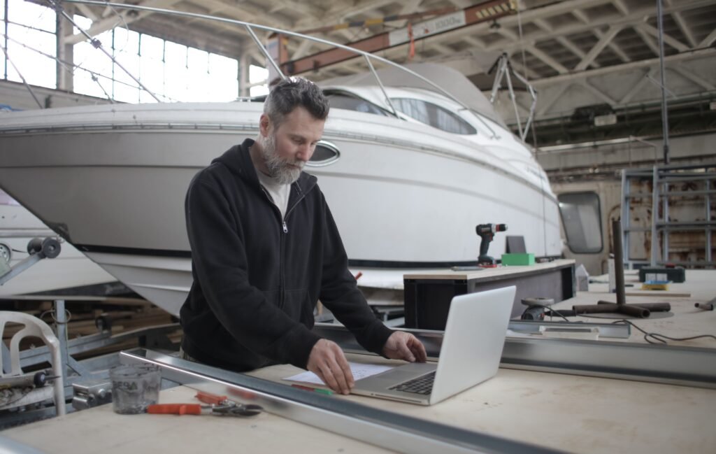 Concentrated adult bearded mechanic in casual outfit using laptop while working with metal details near boat in workshop